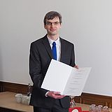 Person mit einer Urkunde steht neben zwei anderen Personen