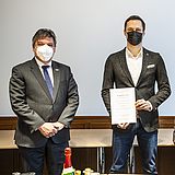 Eine Person hält eine Bescheinigung in der Hand und steht zwei anderen