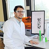 Eine Person mit Laborkittel und Schutzbrille sitzt vor einem Monitor