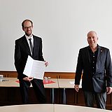 Person mit einer Urkunde steht neben zwei anderen Personen