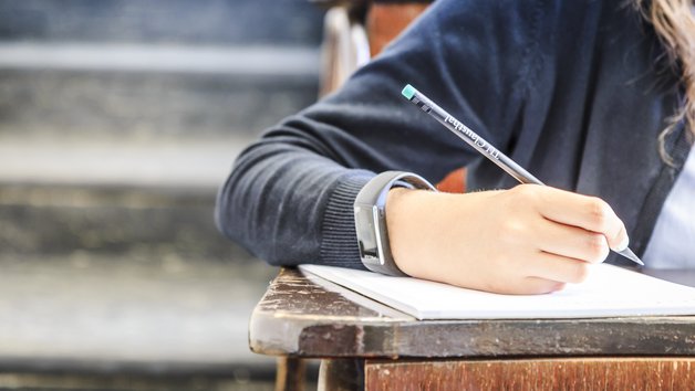 Eine Person schreibt mit einem Stift auf einen Zettel, im Hörsaal sitzend